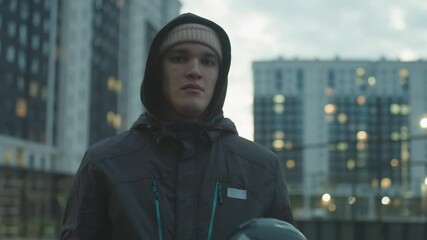 Bored young man in hoodie and beanie juggling football during urban training session, showing casual expression against backdrop of tall residential buildings and cloudy sky