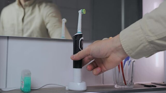 Man removes electric toothbrush from charge and brushes teeth before work in morning. Male in shirt picks up the ultrasonic dental brush view of reflection in mirror. Dental cleaner power tool. 