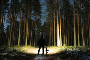 Silhouette of a man with a flashlight and a rifle in a night pine forest on a road in winter.