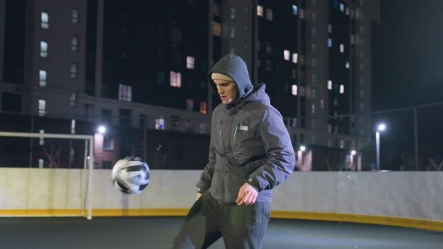 Coach in hoodie joggling football with focus and precision during nighttime training on outdoor field, illuminated by urban lights and surrounded by high-rise buildings and fences