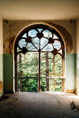 ornate vintage window in abandoned mansion with peeling paint and overgrown trees visible outside,...