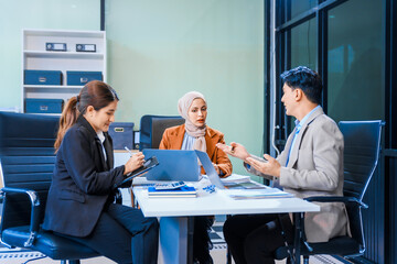 An Asian middle-aged man, a middle-aged Muslim woman in hijab, middle-aged Asian woman collaborate during business meeting. They sit at desk, reviewing report documents and discussing teamwork.