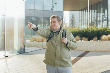 Young 30s blonde man with backpack smiling confident pointing with finger at street. Urban...