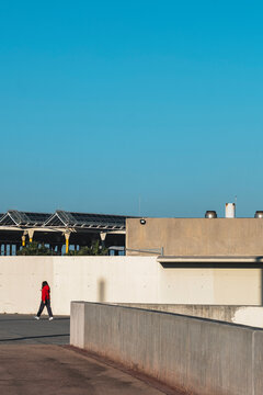 Minimalist urban scene with a single figure walking along a concrete pathway under a clear blue sky. Strong architectural elements and open space define the composition.