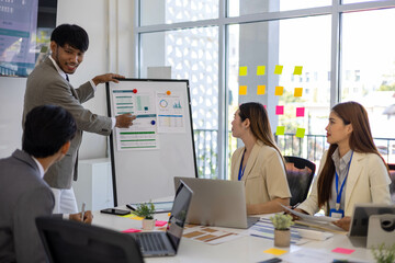 A group of people are working together in a conference room. They are all smiling and seem to be enjoying their time. There are several laptops and papers on the table, and a TV is mounted on the wall