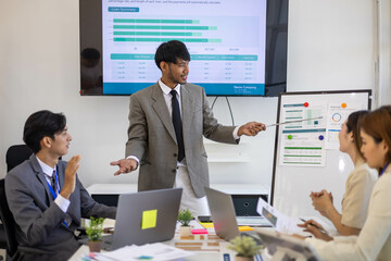A group of people are working together in a conference room. They are all smiling and seem to be enjoying their time. There are several laptops and papers on the table, and a TV is mounted on the wall
