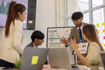 A group of people are working together in a conference room. They are all smiling and seem to be enjoying their time. There are several laptops and papers on the table, and a TV is mounted on the wall