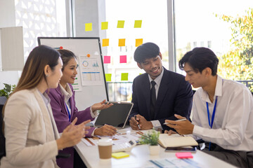 A group of people are working together in a conference room. They are all smiling and seem to be enjoying their time. There are several laptops and papers on the table, and a TV is mounted on the wall