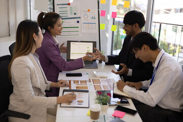 A group of people are working together in a conference room. They are all smiling and seem to be enjoying their time. There are several laptops and papers on the table, and a TV is mounted on the wall