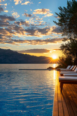 Naklejka premium stunning infinity pool at sunset, featuring wooden deck and lounge chairs, surrounded by mountains and serene lake. vibrant sky enhances tranquil atmosphere