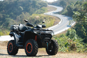 A sleek black ATV parked on a rugged terrain, overlooking a winding road surrounded by lush greenery.