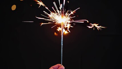 Close-up video of a hand holding a lit sparkler against a dark background, capturing bright sparks from a low-angle perspective.