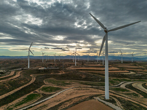 Wind farm in Zaragoza, Spain, captured from above at dusk, featuring towering wind turbines surrounded by intricate roads and fields. The sustainable energy source integrates into nature.