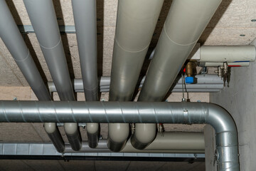 Many pipes for heating and warm and cold water with insulation below the ceiling of the basement of an apartment building.