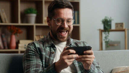 Bearded Caucasian gamer gadget addict man male guy play video game playing online mobile phone smartphone success yelling win internet winning gaming having fun at home couch sofa device entertainment