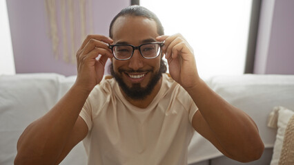 Young man with beard smiling indoors adjusting glasses in cozy living room setting showcasing...