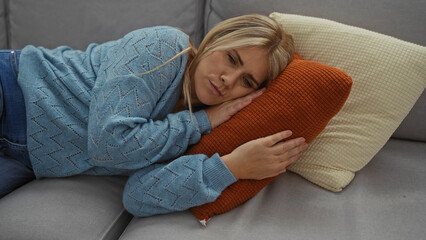 Obraz premium Woman relaxing on a couch in a cozy living room with a blue sweater and blonde hair, resting her head on a soft orange pillow in a serene home environment.