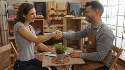 Woman and man shaking hands in a cozy bakery, surrounded by shelves filled with bread and pastries,...