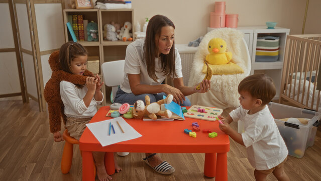 Woman in bedroom offers a banana to a young boy near table with toys, while a girl in brown costume sits nearby, highlighting family bond in playful home setting.