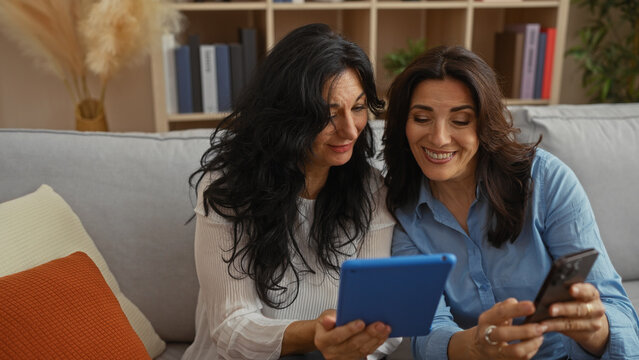 Two mature women sitting on a sofa sharing a moment of connection with digital devices in a cozy living room environment, highlighting friendship and technology use.