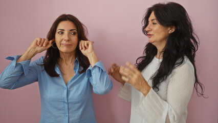 Middle-aged sisters engaging in a playful conversation, one covering her ears, set against a pink background expressing love and family connection.