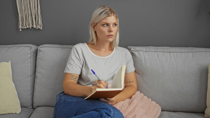 Young woman with blonde hair writing in a notebook sitting on a couch in a living room, appearing...