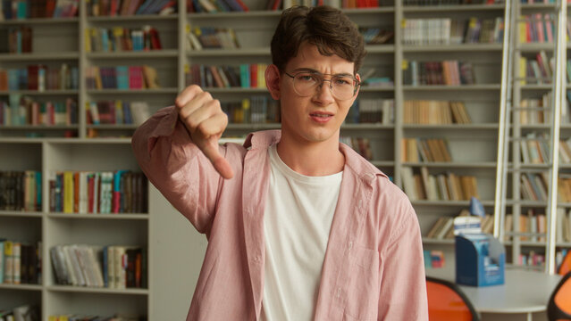 In school library book store university college upset frustrated Caucasian student boy looking at camera dissatisfied man male schoolboy guy education studying showing dislike thumb down no answer