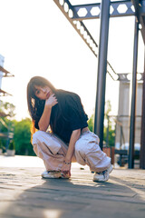 Young woman squatting in casual outfit under warm sunlight in outdoor urban setting