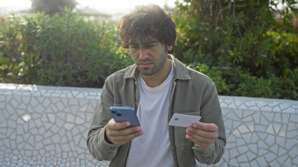 Young man outdoors using smartphone and credit card in urban park setting, focused on online banking or shopping, surrounded by greenery and enjoying a sunny day.