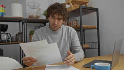 Fototapeta premium Man reading documents at desk in modern office with laptop and coffee, displaying focused expression and casual attire, surrounded by shelves and kitchen utilities.