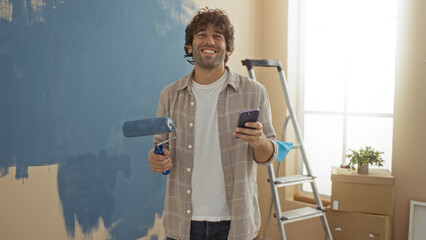 Young man painting wall with roller in new home while checking phone in brightly lit living room...