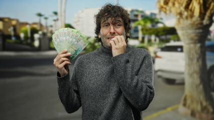 Hispanic man nervously biting nails on city street holds russian roubles, conveying anxiety around...