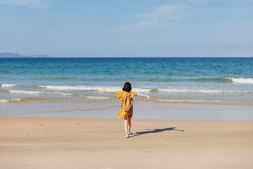 Woman in yellow dress walking on beach with arms outstretched enjoying the beauty of nature and feeling free