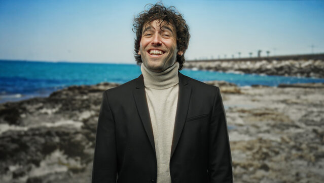 Handsome young man smiling confidently at seaside with blue sky and ocean waves, wearing elegant jacket, embodying joy and relaxation on a peaceful sunny beach day