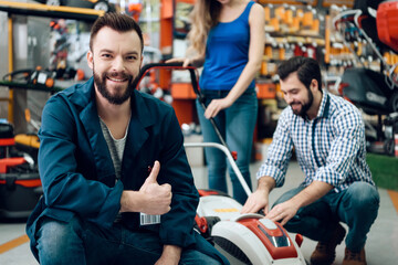 Salesman in blue robe is posing with couple of clients and new lawn mover in power tools store.