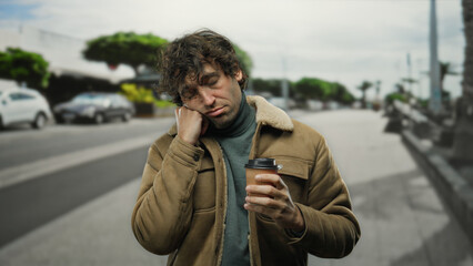 Obraz premium Young man outdoors on city street looking sleepy and holding coffee, wearing jacket, with cars in background and trees lining the road, displaying urban lifestyle.