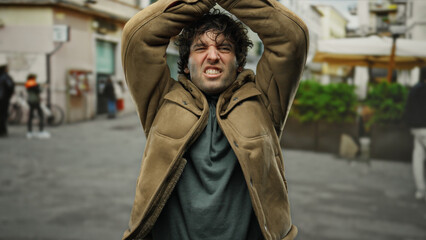 Young hispanic man looking frustrated outdoors on city street in casual jacket with arms raised, capturing an intense emotion in an urban setting.