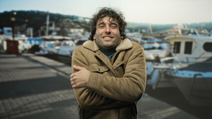 Fototapeta premium Young man outdoors at port feeling cold amidst boats, wearing a jacket, handsome, hispanic, attractive, seaside setting, showing shivering gesture.