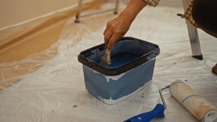 Woman painting in new home with paint bucket and brush on tarp covered floor creating a fresh and...