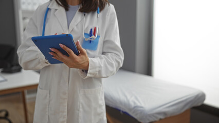 Female doctor holding a tablet in a clinic room with a stethoscope around her neck, showcasing a professional healthcare environment indoors.