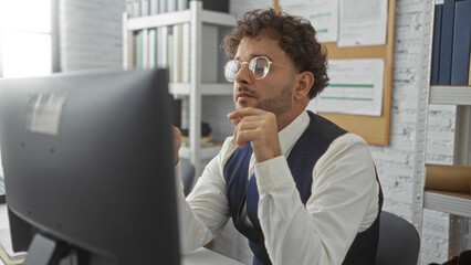 Young hispanic man working intently at a computer in an office setting with bookshelves and documents around him showing focus and professionalism.
