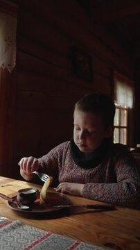 Vertical video of preteen boy eating fried potato pancakes with sour cream in cafe. Child dips national Slavic dish draniki into gravy boat and eats.