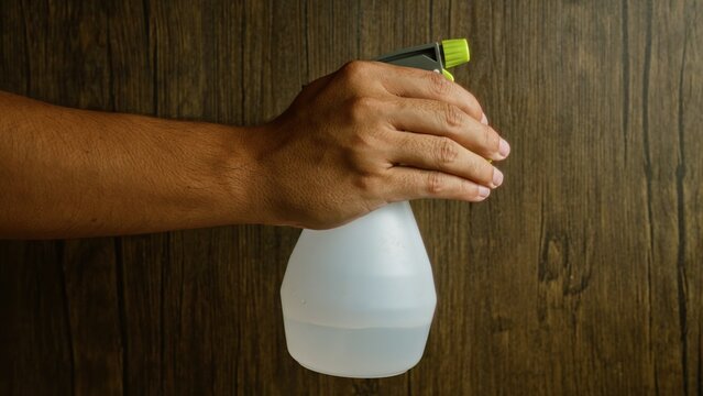Man holding white spray bottle against wooden background, displaying a cleaning or gardening tool in a simple setting, emphasizing the hand detail and texture of the wood surface