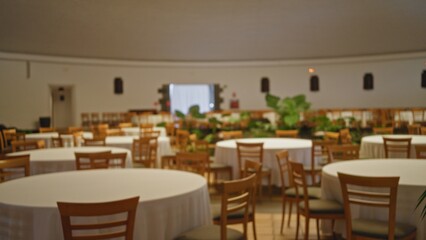 Blurred image of an empty wedding hall with round tables draped in white tablecloths and wooden chairs, featuring a defocused background with plants and softly lit decorations.