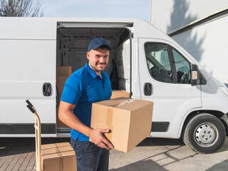 Smiling delivery worker carrying a cardboard box. Courier in a blue uniform smiling while carrying a cardboard box next to his delivery van. Logistics and shipping concept.