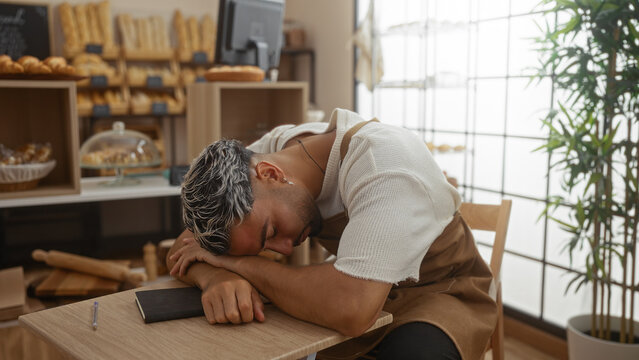 Tired man sleeping at a table in a bakery shop during working hours, surrounded by bread and pastries