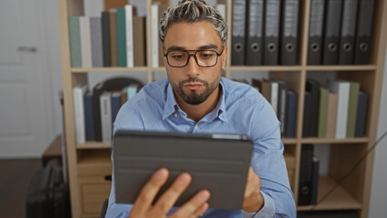 Young, arabian, bearded, man reading tablet in indoor office setting with bookshelves in the...