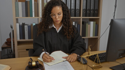 Hispanic woman with curly hair in a judge's robe writing in a notebook in an office with a...