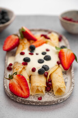 Crepes, blinis or thin pancakes with strawberries, whipped cream and blueberries on a plate on stone table. Sweet Breakfast or Brunch. Maslenitsa, Mardi grass traditional meal.