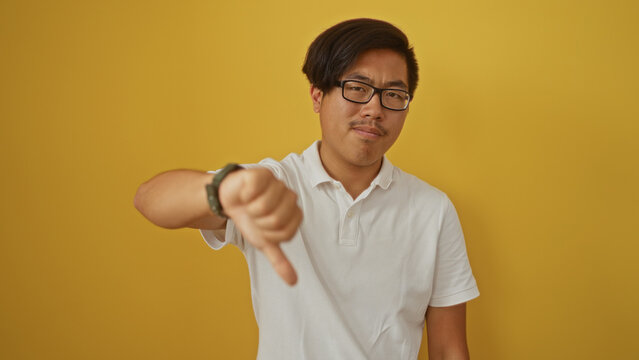 Young man giving a thumbs down gesture against a yellow background wearing a white shirt, displaying dissatisfaction.